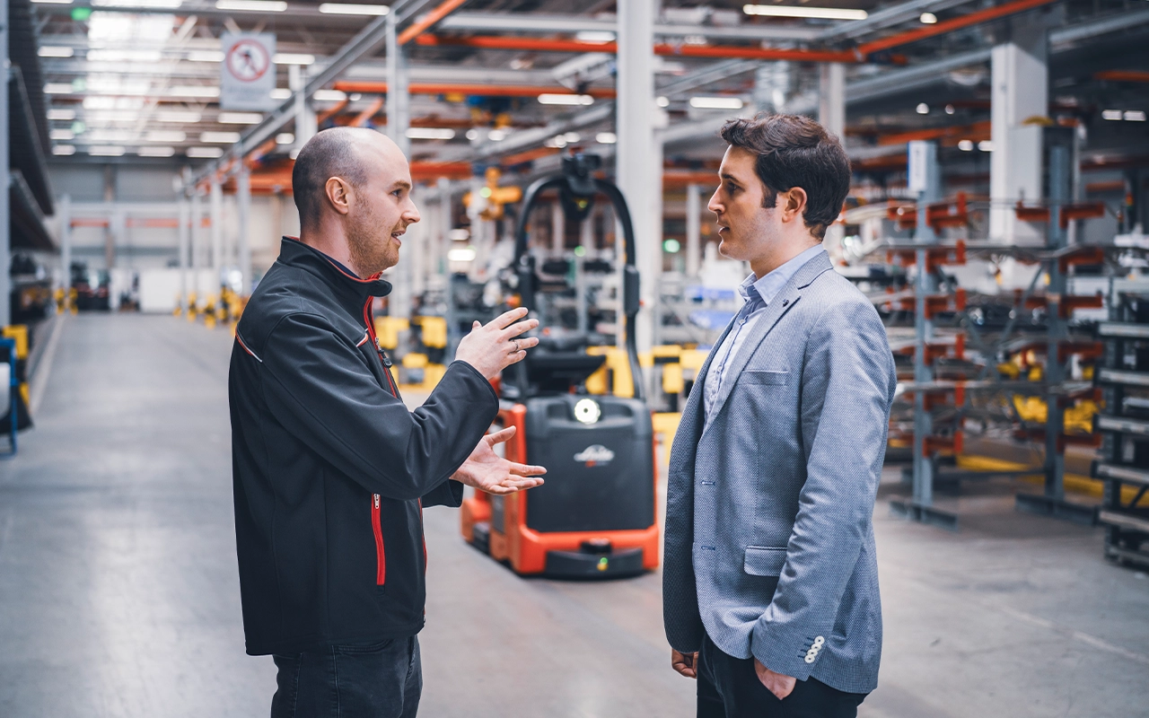 Ein Linde-Mitarbeiter in schwarzer Jacke spricht in einer Produktionshalle mit einem Mann im grauen Sakko. Hinter ihnen steht ein roter Linde-Hubwagen.