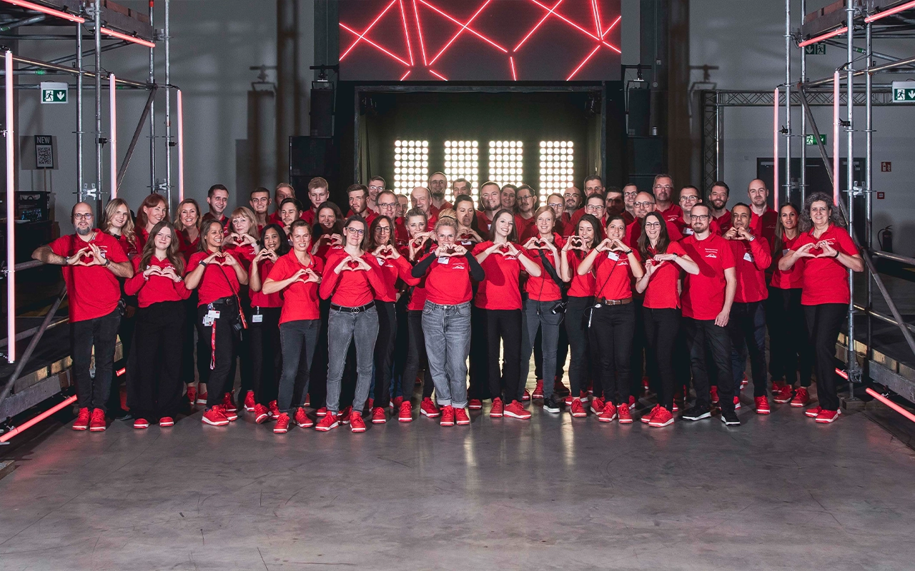 Gruppenfoto eines großen Teams in einheitlich roten Shirts und roten Sneakern in einer Veranstaltungshalle. Alle Personen formen ein Herz mit ihen Händen.
