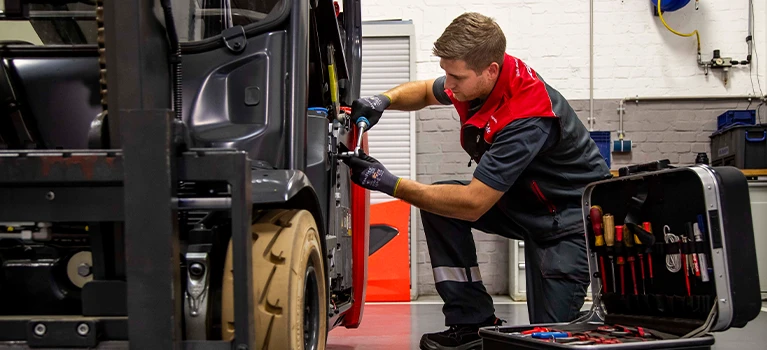 Ein Servicetechniker in rot-grauer Arbeitskleidung kniet in einer Werkstatt neben einem Gabelstapler und repariert ihn. Neben ihm steht ein geöffneter Werkzeugkoffer.