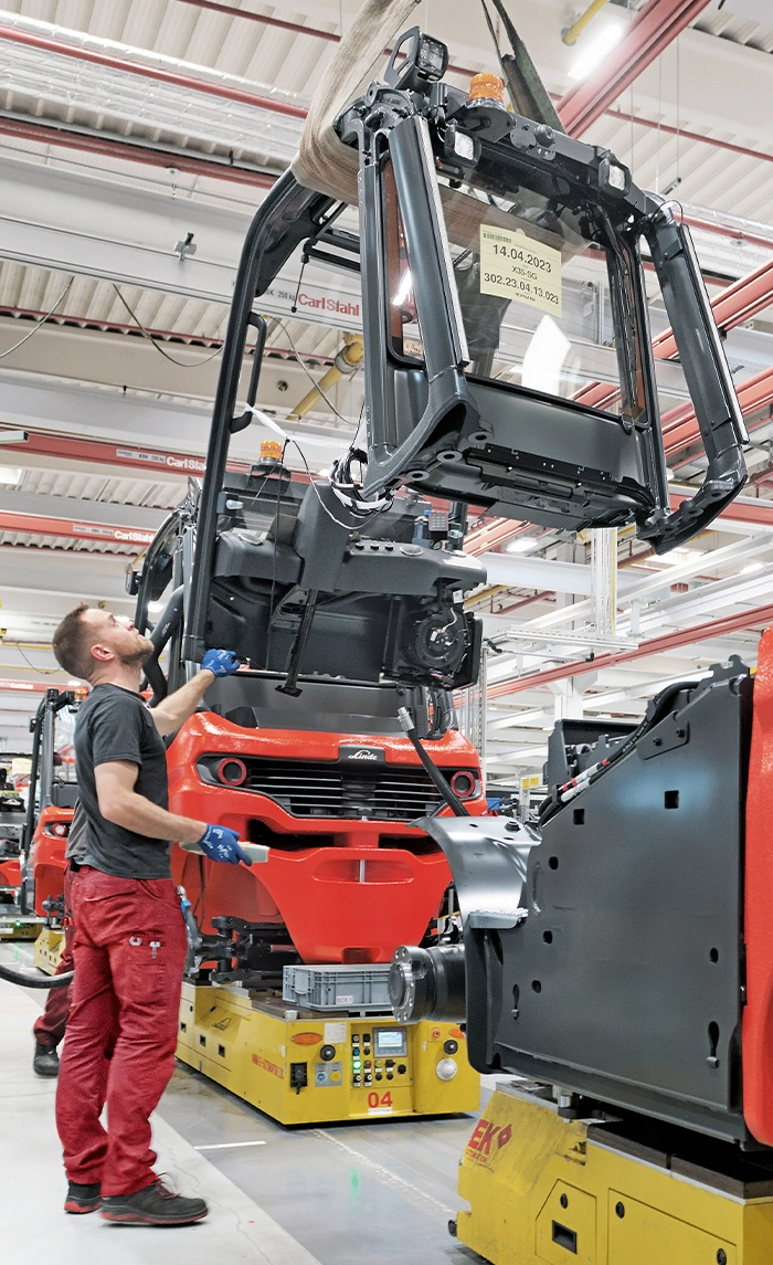 Ein Mitarbeiter an der Fertigungslinie führt eine am Kran hängende Fahrerkabine auf einen roten Linde-Gabelstapler, der auf einer automatisierten Transportplattform steht.