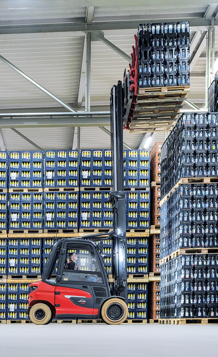 Ein roter Linde-Gabelstapler hebt in einem Lager voller gestapelter Getränkekisten mehrere Paletten mit Kisten weit nach oben.