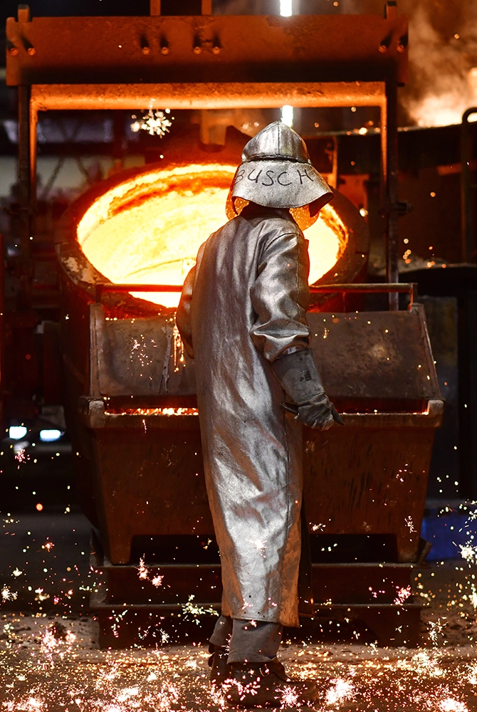 Ein Gießereiarbeiter im silbernen Hitzeschutzanzug steht vor einer Pfanne mit glühendem, flüssigem Metall, während Funken über den Boden sprühen.