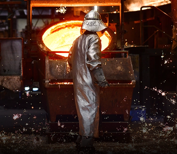 Ein Gießereiarbeiter im silbernen Hitzeschutzanzug steht vor einer Pfanne mit glühendem, flüssigem Metall, während Funken über den Boden sprühen.