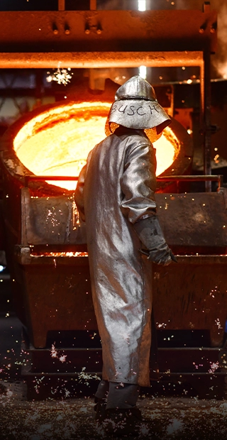 Ein Gießereiarbeiter im silbernen Hitzeschutzanzug steht vor einer Pfanne mit glühendem, flüssigem Metall, während Funken über den Boden sprühen.