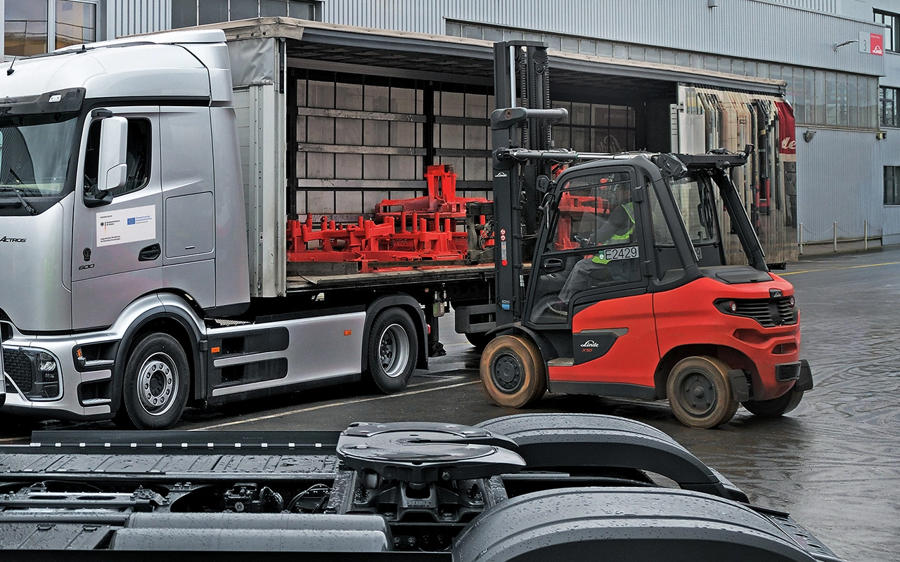 Ein roter Linde-Gabelstapler belädt vor einer Werkshalle einen Lkw mit geöffneter Plane, auf dessen Ladefläche rote Transportgestelle stehen.