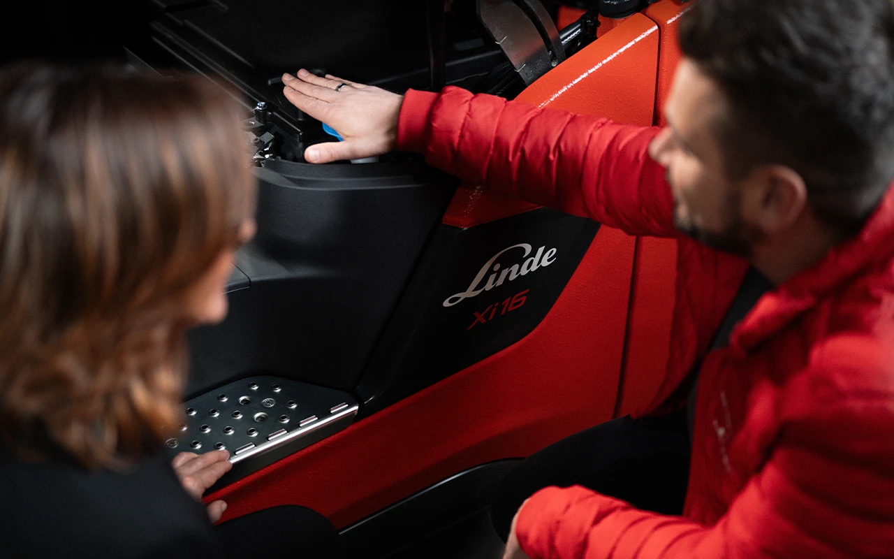 A service technician in a red jacket shows a woman the battery compartment of a red Linde Xi16 electric forklift.