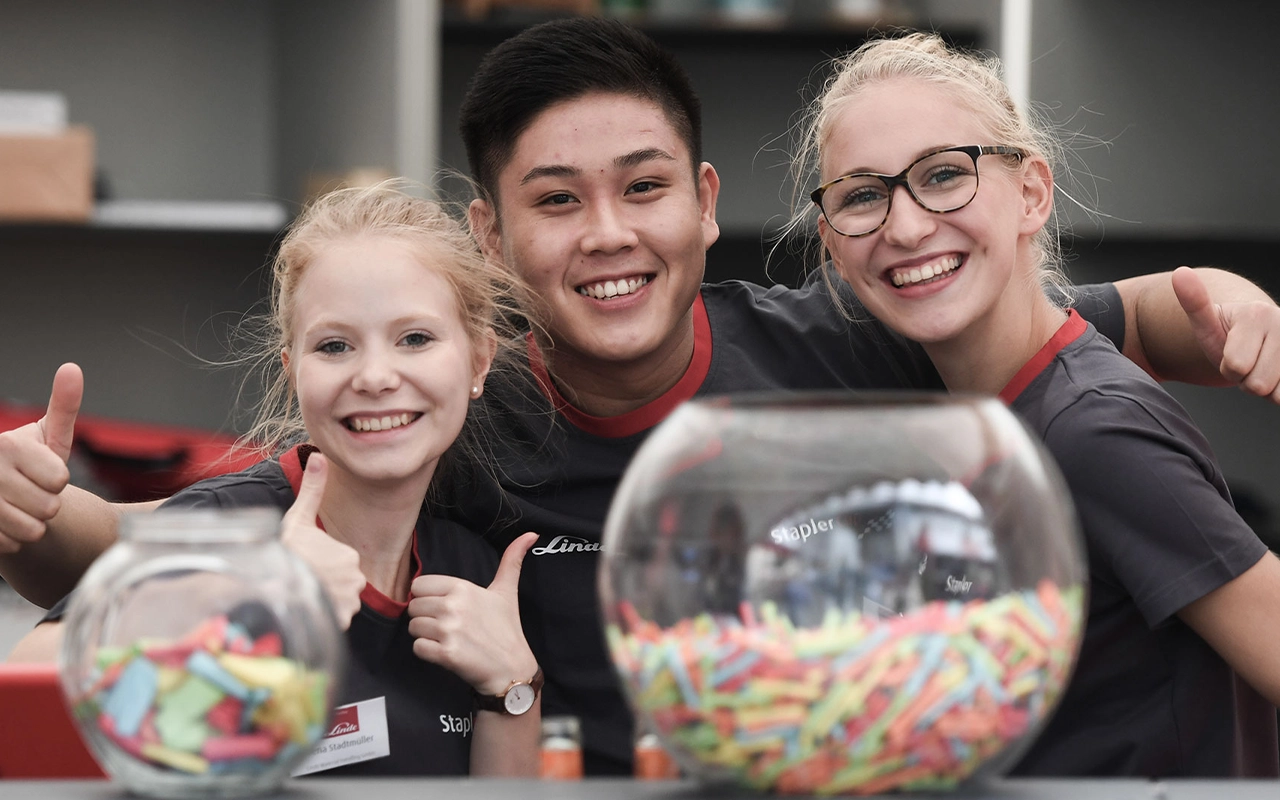 Three smiling young Linde employees in branded shirts. Two of them are giving a thumbs-up with both hands. In front of them are two bowls with colourful contents.
