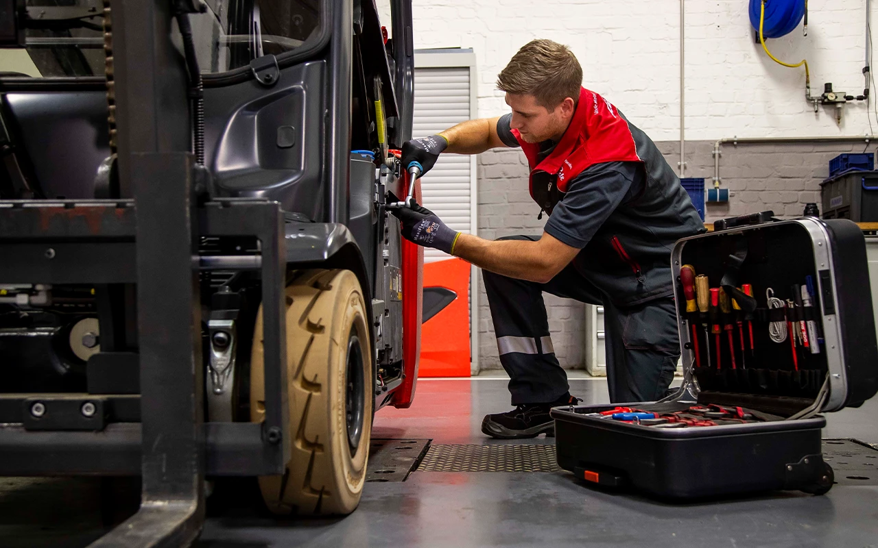 A service technician in a red and grey uniform kneels beside a forklift in a workshop and repairs it, with an open toolbox next to him.