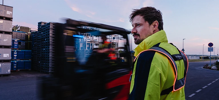 A man wearing a yellow high-visibility jacket stands on an outdoor prouction yard at dusk while a red forklift drives past him in a motion blur.