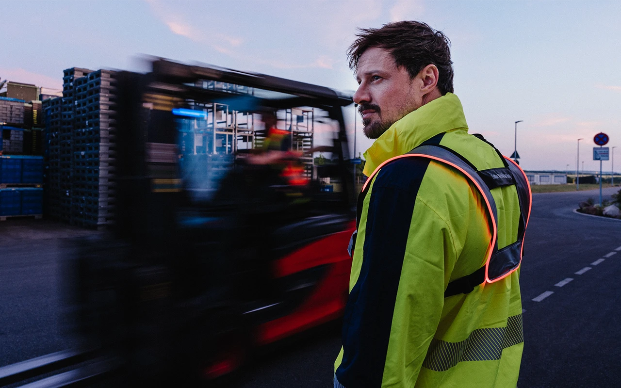 A man wearing a yellow high-visibility jacket stands on an outdoor prouction yard at dusk while a red forklift drives past him in a motion blur.