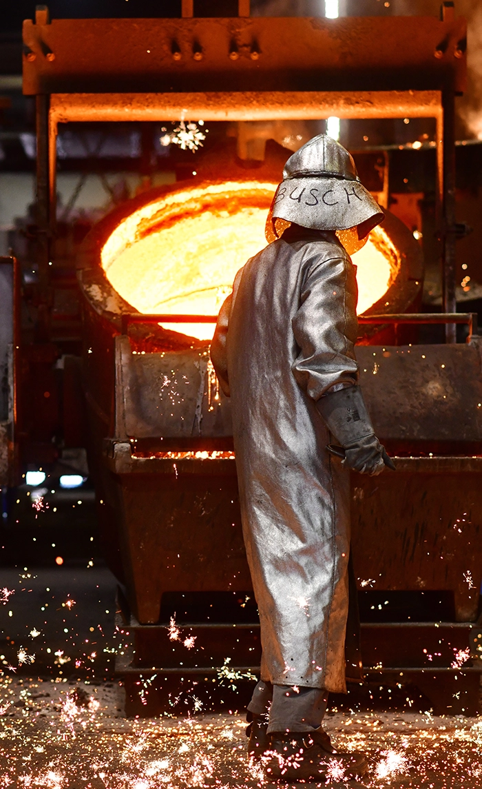 A foundry worker in a silver heat-protective suit stands in front of a ladle of glowing molten metal as sparks fly across the floor.