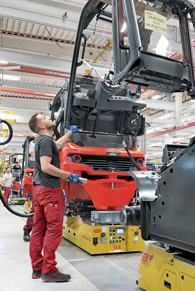 A worker on a production line guides an overhead guard, suspended from a crane, onto a red Linde forklift on an automated transport platform.