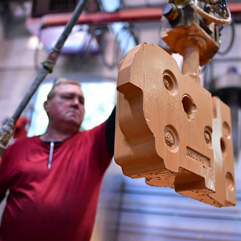 A foundry worker guides an orange sand moulding part held by a lifting device.