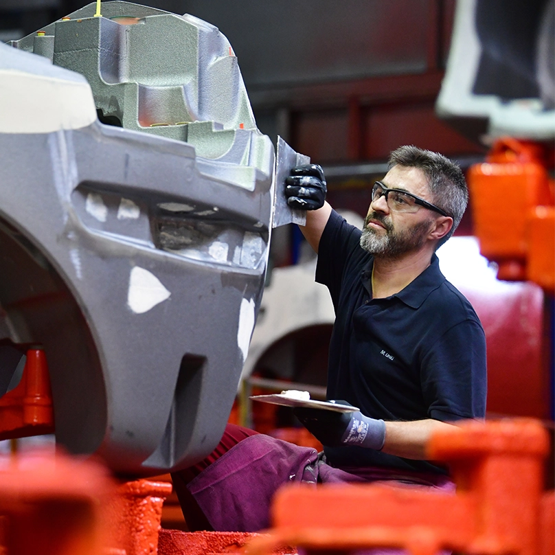 A worker with safety glasses applies filler with a spatula to a large grey cast counterweight for a forklift.