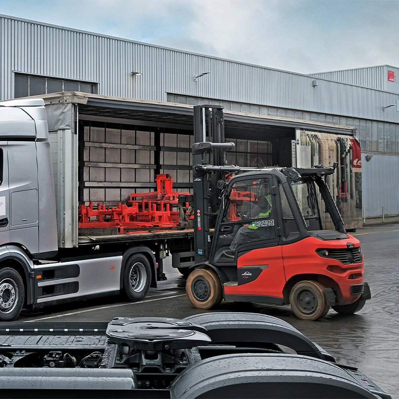 A red Linde forklift loads a truck with an open curtain side at a factory building. There are red tansport racks in the truck. 