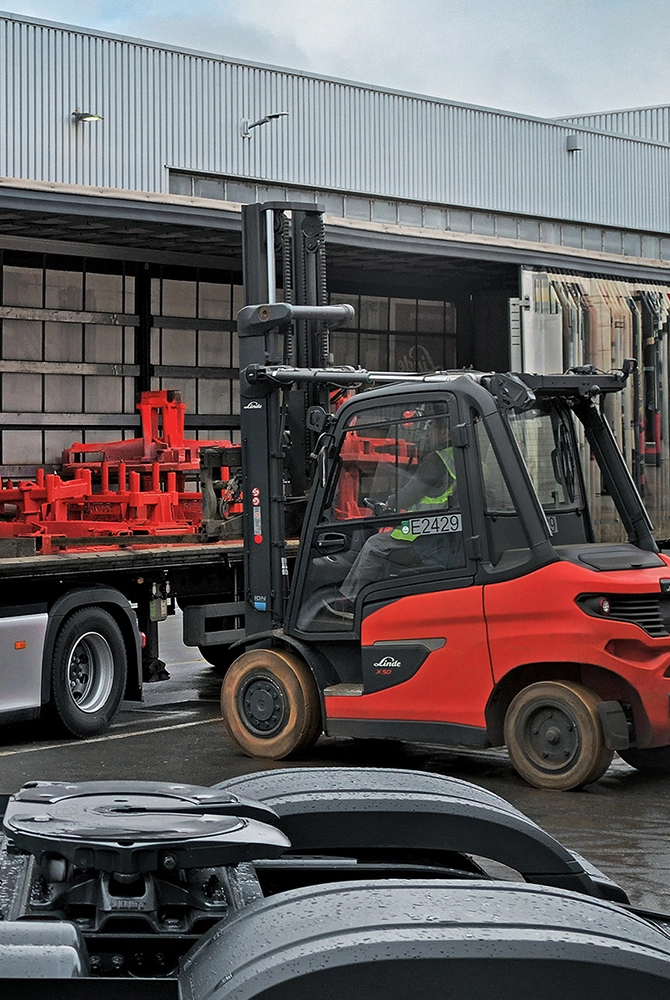 A red Linde forklift loads a truck with an open curtain side at a factory building. There are red tansport racks in the truck. 