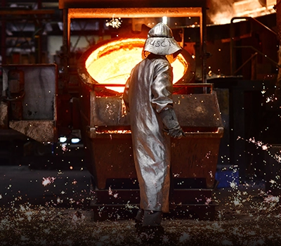 A foundry worker in a silver heat-protective suit stands in front of a ladle of glowing molten metal as sparks fly across the floor.