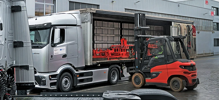 A red Linde forklift loads a truck with an open curtain side at a factory building. There are red tansport racks in the truck.