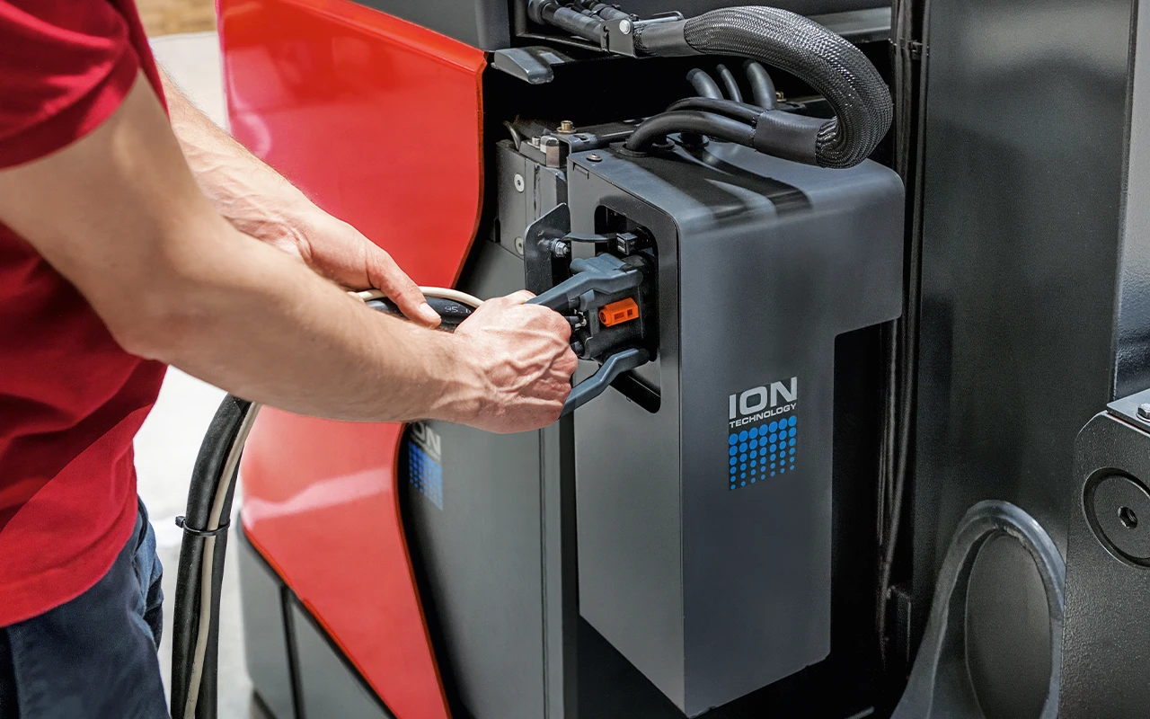 A man plugs a charging cable into a battery labelled "ION Technology" on a red industrial truck.